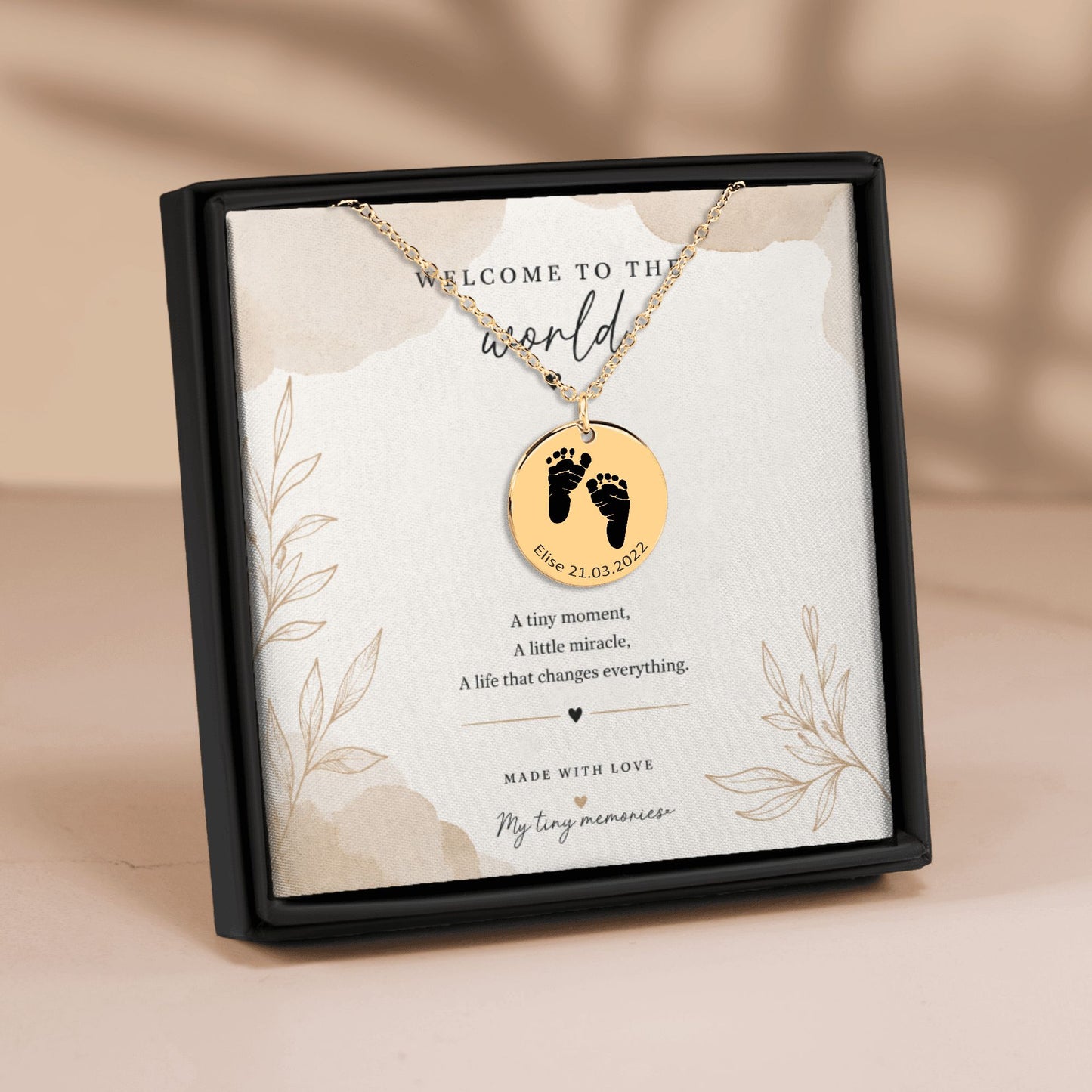 Necklace with a round pendant featuring footprints and text on a branded card, set against a beige background.