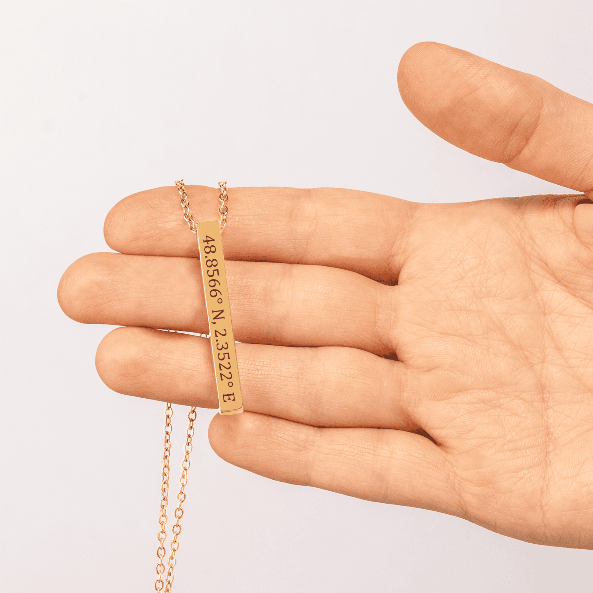 Gold necklace with engraved coordinates held between fingers on a light background
