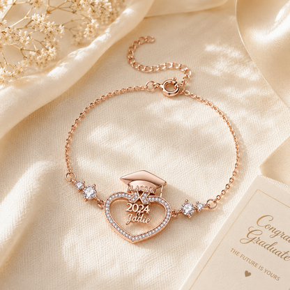 Rose gold bracelet with heart-shaped graduation cap and diploma charm on a beige fabric background