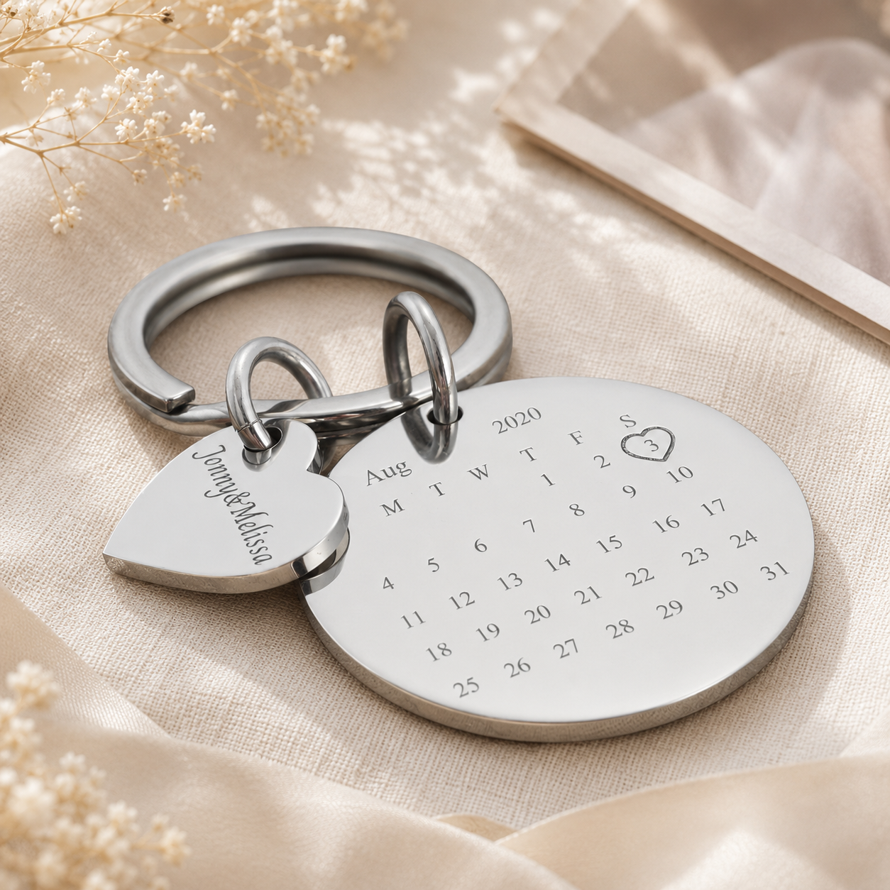 Silver keychain with heart-shaped tag and calendar engraving on a beige fabric background